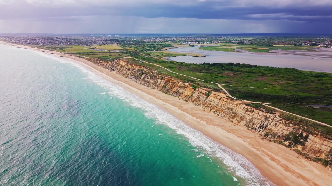 Droneoptagelse langs Hengistbury Head kystlinje med turkis hav, sandstrand, kystklipper, hede, havn, vådområder og dramatisk skyet himmel.