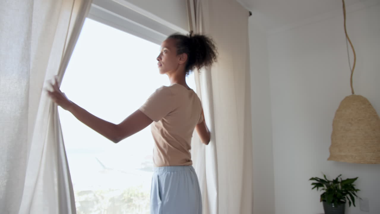 Donna che apre le tende a casa, guardando pensierosa fuori dalla finestra
