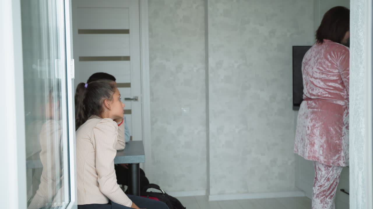 Girl and boy sit silently at table in corner while woman in pink velvet outfit prepares food at kitchen counter, showing quiet family routine, emotional distance, and contrast between roles in home setting