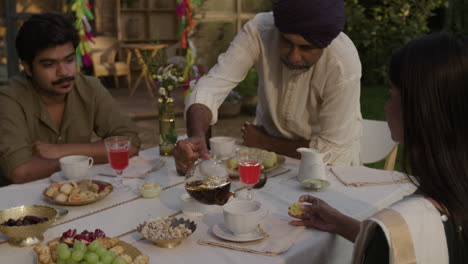 family gathering outdoors with a man pouring tea at a table filled with snacks