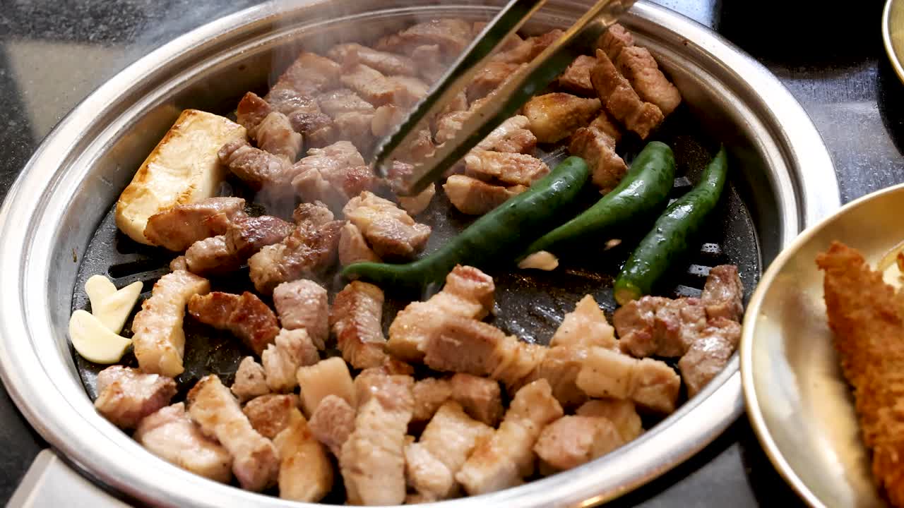 Chopsticks turn sizzling pork belly, peppers, garlic, and tofu on a tabletop Korean barbecue grill