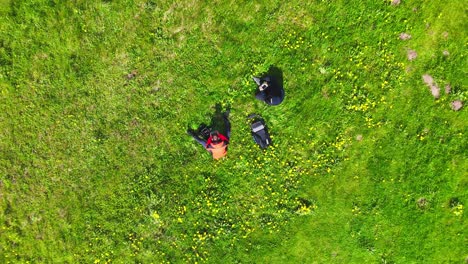 two drone pilots stand in a green field operating quadcopters as gear rests nearby, captured in a clean overhead frame that suggests instruction, teamwork, and safe recreational flight