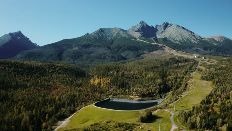 Aerial-drone-shot-of-the-High-Tatras-forest-with-a-geomembrane-water-dam-and-cableway,-showcasing-green-summer-trees,-hills,-and-mountains-in-Slovakia,-Europe