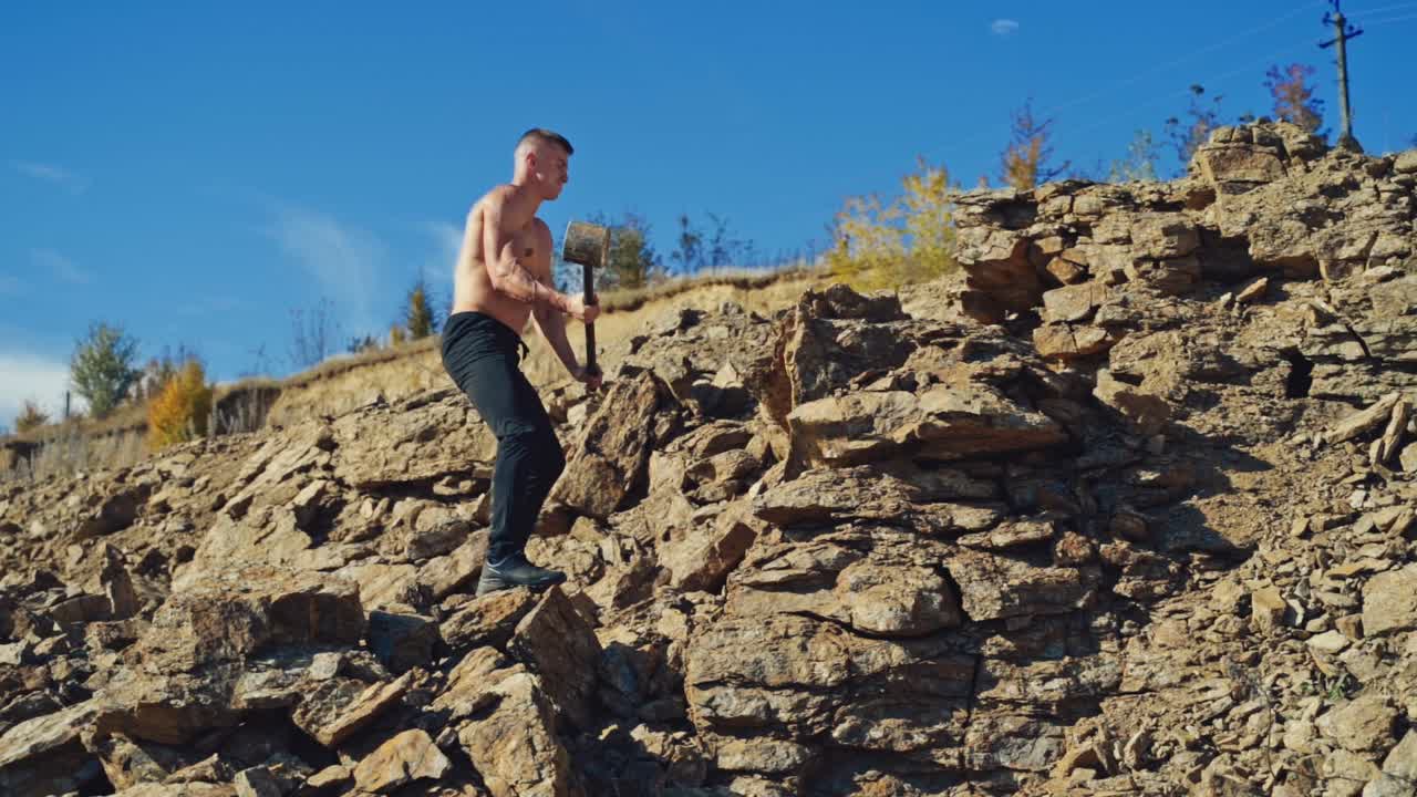 Side view of a strong man breaking stones with a hammer. Shirtless athletic man exercising with a sledgehammer on the rocky background. Slow motion.