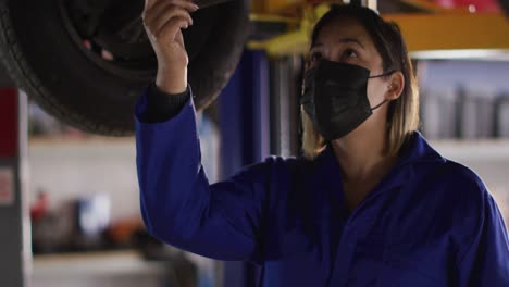 female mechanic wearing face mask holding digital tablet inspecting the car at a car service station