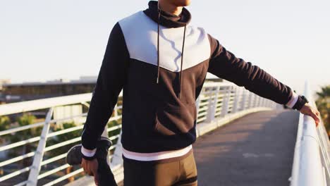 african american man wearing earphones exercising outdoors, standing on footbridge stretching leg