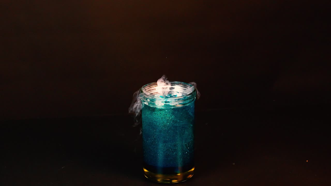 A beaker with blue liquid and dry ice creates a smoky, bubbling effect in a dark setting