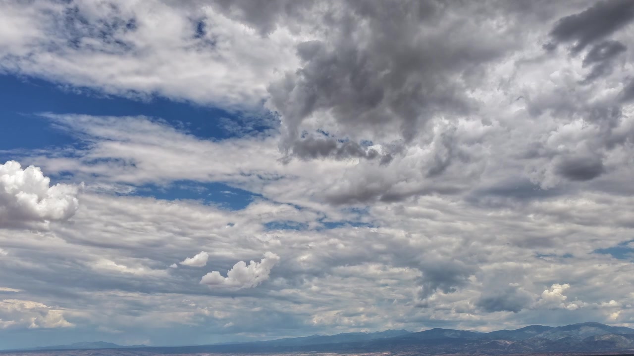 4K aerial timelapse capturing dramatic layers of clouds shifting rapidly across a deep blue sky. The fast-moving weather system rolls over the distant, rugged peaks of Mt Baldy in the background.
