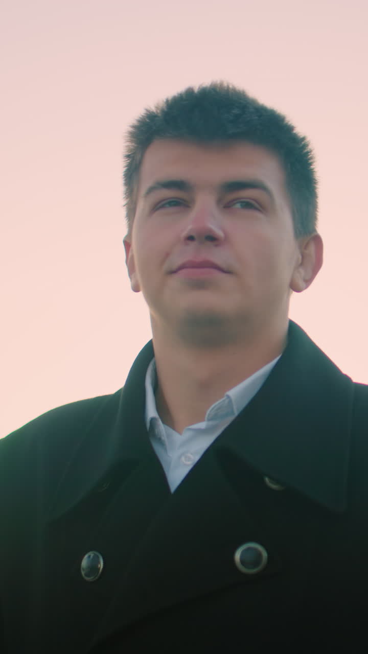 사업가 검은 슈트 야외에서 황혼에 자신감 있는 표정으로 커피를 마시고, 배경에는 현대적인 건물, 주차된 자동차 및 구조물이 있습니다.