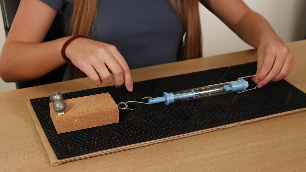 A person conducts a friction experiment using a spring scale and weights on a textured surface, illustrating force measurement techniques