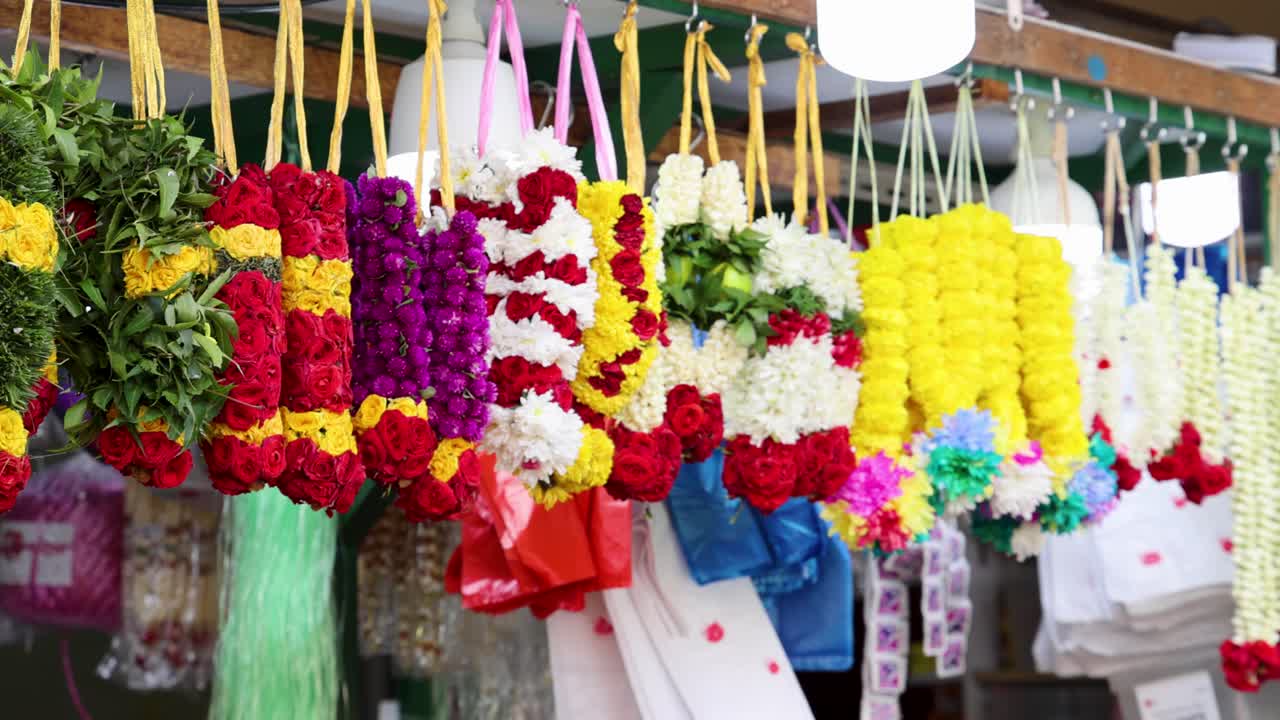 Rows of vibrant flower garlands, including marigold and rose, hang from a market stall in Little India, Singapore. Bright daylight, slow camera pan