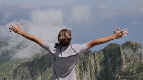 young man stands on mountain top above clouds, arms wide open.