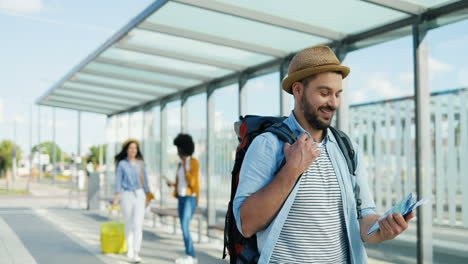 Young-handsome-Caucasian-traveller-wearing-hat-with-backpack-holding-city-map-while-walking-at-bus-station