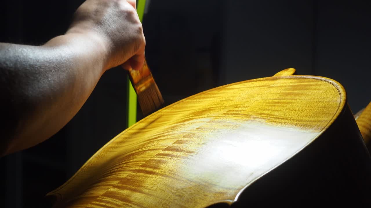 Close-up of a craftsman’s hand spreading a homemade mix of linseed oil, mastic, and lac on the cello’s marbled back, with vivid wood flames revealed by focused strong studio lighting