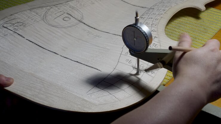 Maestro liutaio, costruttore di strumenti a corda, procede con la lavorazione di graduazione sulla tavola posteriore di un violoncello, utilizzando un calibro di spessore per misurare i livelli e una matita per annotare i valori sul legno, nel suo laboratorio a Cremona, Italia.