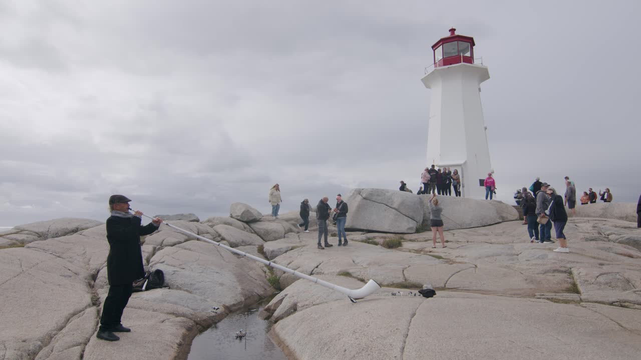 Tourists gather at Peggy's Cove lighthouse as a musician plays an alpenhorn on rocky terrain