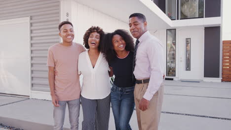 black family stand looking to camera in front of their modern home, three quarter length