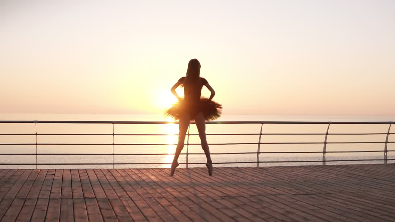 Young, tender ballet dancer practicing near the sea ocean. Jumping up on the spot. In dark tutu and pointe. Backside view. Morning