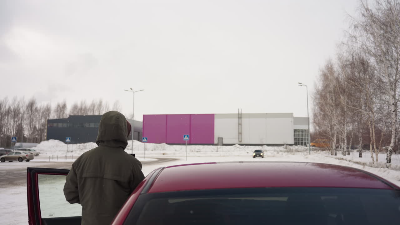 rear view of driver in winter hoodie standing by open car door on snowy roadside, distant car approaching on snow-covered road with urban buildings and trees in background during cold overcast day