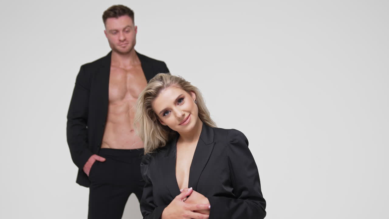 Beautiful long-haired blonde woman stands on her knees waving her hair. Man in black suit stands at backdrop. White background.