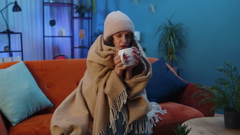 sick unhealthy woman wear hat wrapped in plaid sit, shivering from cold on sofa drinking hot tea