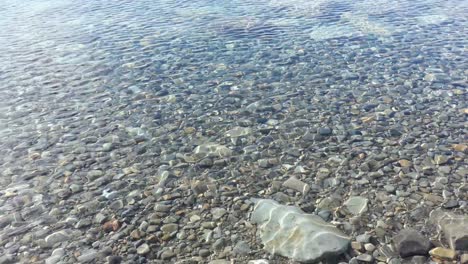 Slow-walk-beside-beautiful,-clear-water-of-alpine-lake-showing-vast-array-of-stones-and-colors