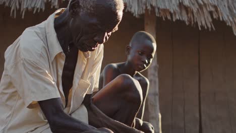 elderly man sits outside a thatched roof hut, meticulously peeling food, while a young child sits beside him, observing the process in a rural village setting