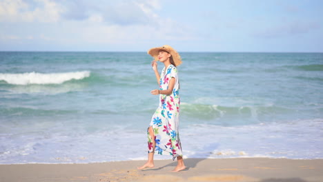 slow-motion of asian lady walking along the beach in thailand wearing a long sundress, she is holding her straw hat because its windy day, slow-motion following show side view