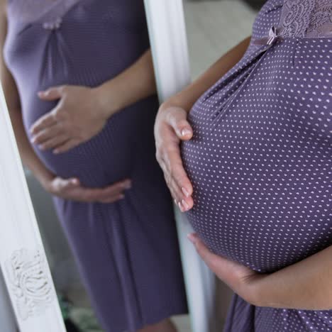 woman on the last months of pregnancy standing in front of the mirror. pregnant lady in violet sleepwear watching at her reflection in the mirror