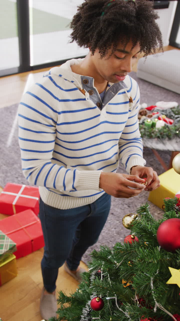 video vertical de un hombre feliz de dos razas con cuernos de reno decorando el árbol de navidad en casa