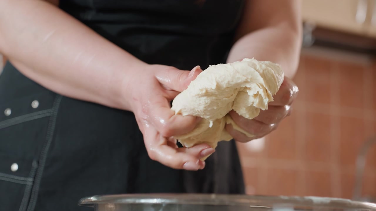 Mani adulte lavorano un impasto appiccicoso sopra una ciotola di metallo in una cucina accogliente, con movimenti tattili decisi, piegando, premendo, allungando e modellando per la preparazione del pane fatto in casa sul piano di lavoro.