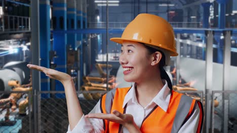 primer plano de una ingeniera asiática con casco de seguridad de pie en una fábrica de fabricación de turbinas eólicas. sonriendo y señalando hacia un lado recomienda algo mientras el brazo robótico trabaja