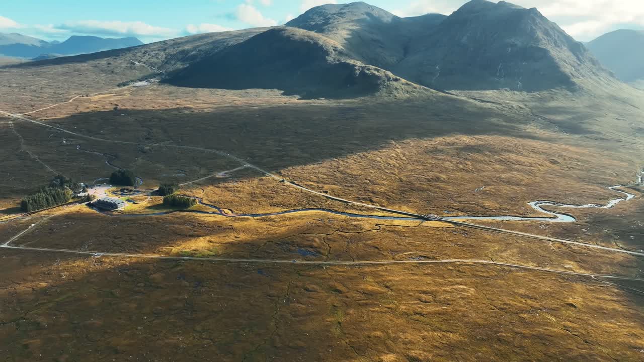 Ripresa aerea in volo sopra la valle di Glencoe, Highlands scozzesi, Scozia, che mostra il paesaggio della brughiera, il sentiero della West Highland Way, il Kings House Hotel e le montagne circostanti in una giornata con cielo azzurro e nuvoloso.