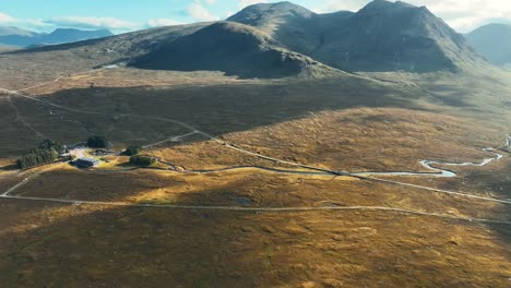 Aerial-shot-flying-over-valley-in-Glencoe,-Scottish-Highlands,-Scotland-viewing-the-moorland-landscape,-West-highland-way-path,-Kings-House-Hotel-and-mountains-surrounding-on-a-blue-sky-and-cloudy-day