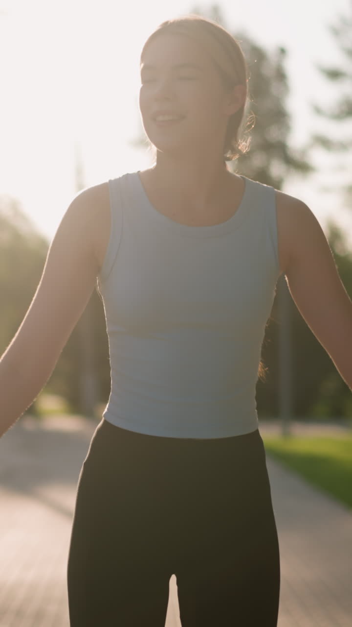 Young lady smiling while skating outdoors, swinging her hands for balance, with sunlight reflecting off her creating a bright glow, blurred background featuring lush greenery and trees