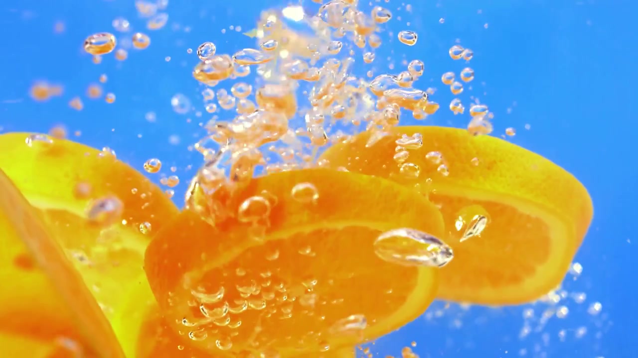 Slow motion orange slices splash into clear water against a bright blue background, creating bubbles and a fresh citrus look for drinks, summer, health, and food ads.
