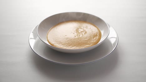 steaming hot butternut squash  soup being poured into a white soup bowl, sitting on a white plate on a white table