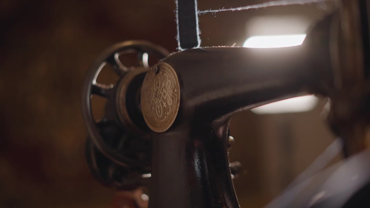 Primo piano di una macchina da cucire antica, mano che guida la ruota del tessuto nella luce calda dell'officina, ago e bobina visibili, focus sulla patina del metallo e sul filo testurizzato, scena di sartoria tranquilla e attenta.