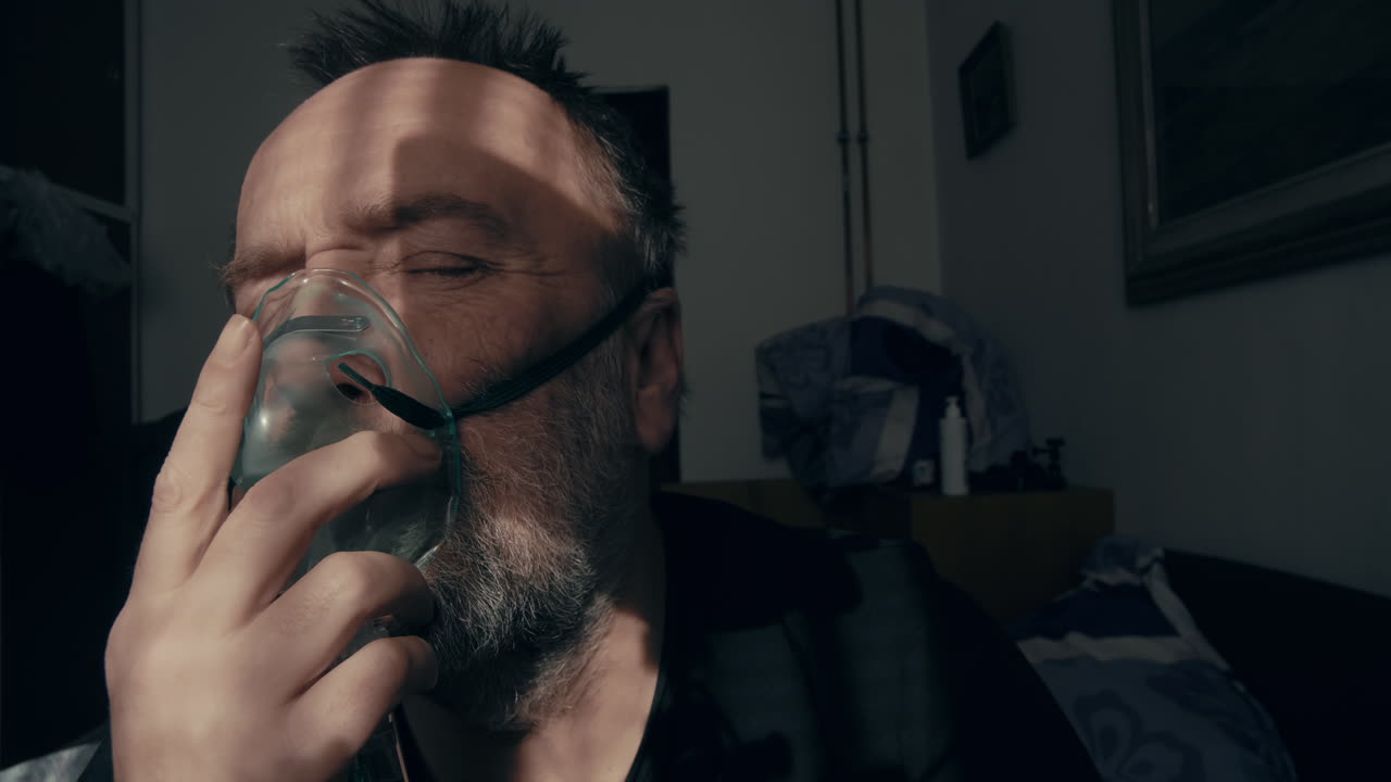 Primo piano di un uomo maturo con la barba che respira attraverso una maschera per l'ossigeno in una stanza poco illuminata. La luce solare che filtra dalle persiane crea ombre drammatiche sul suo viso. Concetto di asma, trattamento BPCO, post-virale.