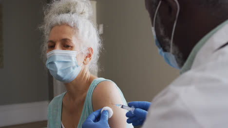 male african american doctor wearing face mask injecting covid-19 vaccine into senior female patient