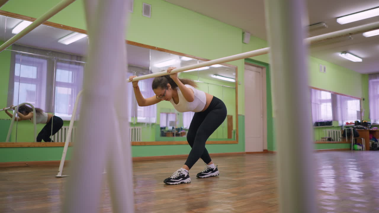Girl in casual wear with sneakers exercising indoors, holding bar while bending forward in studio with green walls, mirrors, and wooden floor, showing focus, strength, and controlled body posture