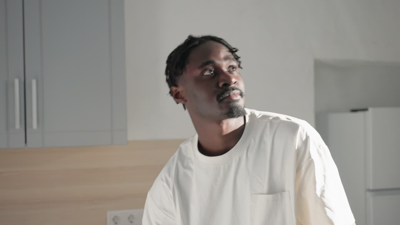 Man wearing casual white shirt and backpack looking up in modern kitchen, with white walls and gray cabinets in the background. Refrigerator and kettle are visible in the kitchen area