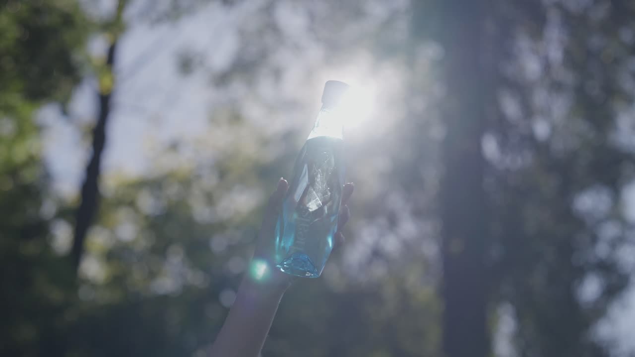 Persona che solleva una borraccia blu di vetro con la luce del sole e gli alberi sfocati