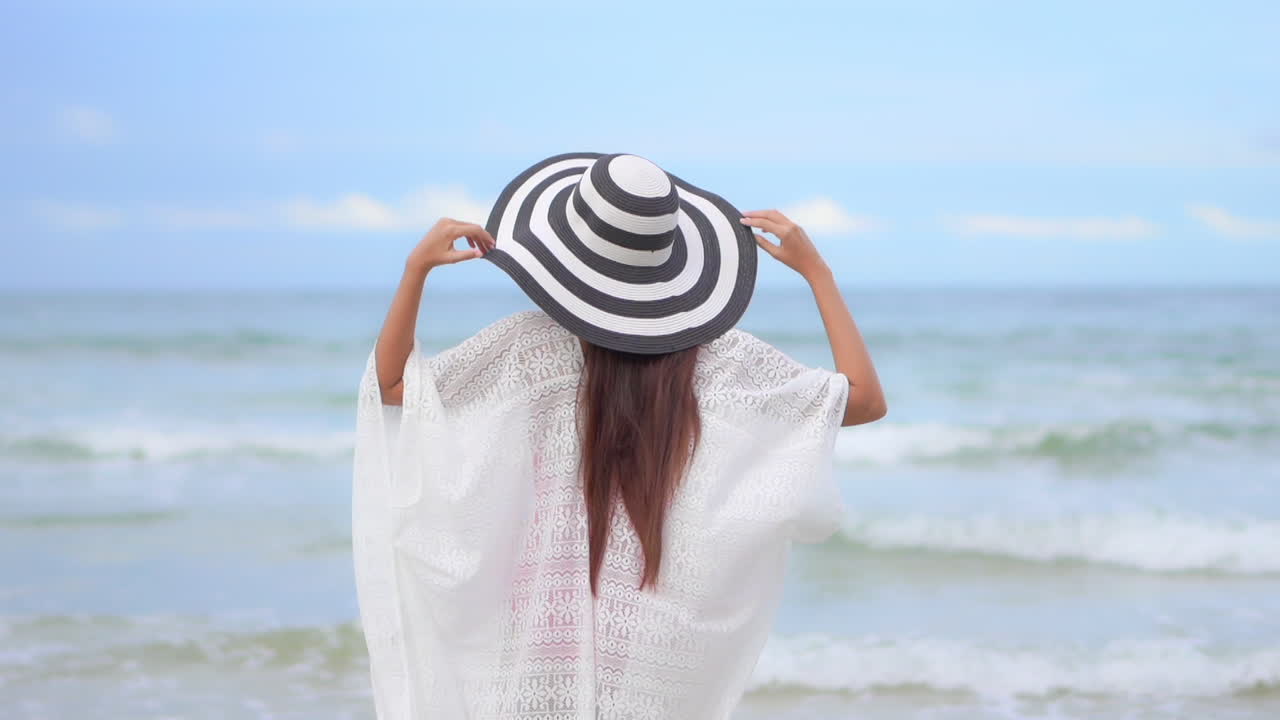 튜닉과 여름 모자를 쓴 품격 있는 여성의 뒷모습, 모래 열대 해변의 파도 앞에서 걷는 모습, 선택적인 초점, 풀 프레임, 느린 동작
