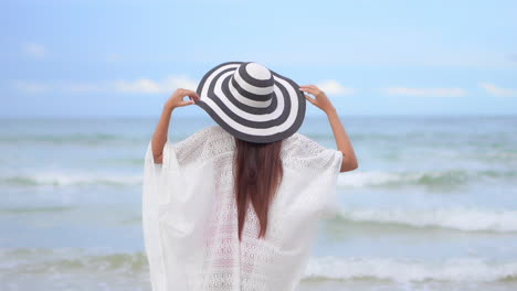 Back-View-of-Classy-Female-in-Tunic-and-Summer-Hat-Walking-in-Front-of-Sea-Waves-on-Sandy-Tropical-Beach,-Selective-Focus,-Full-Frame,-Slow-Motion