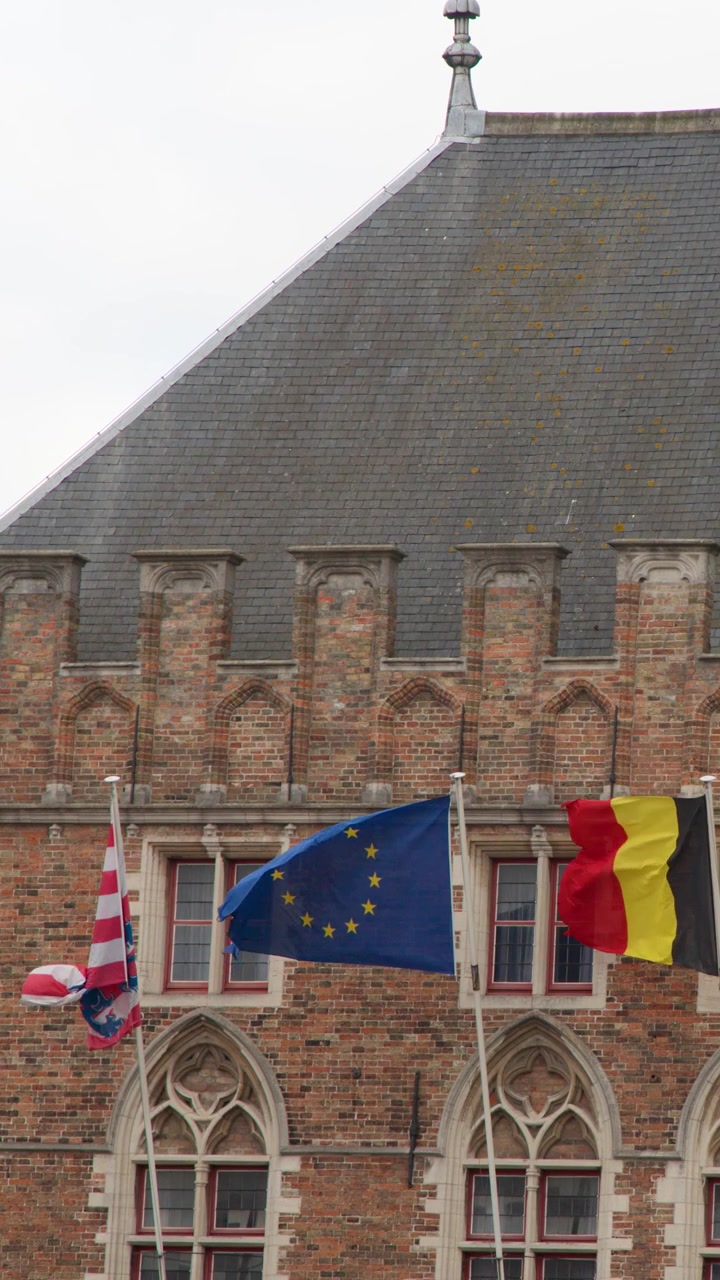 Bandiere dell'UE e del Belgio sventolano su un edificio medievale in mattoni sotto un cielo coperto, ripresa statica.