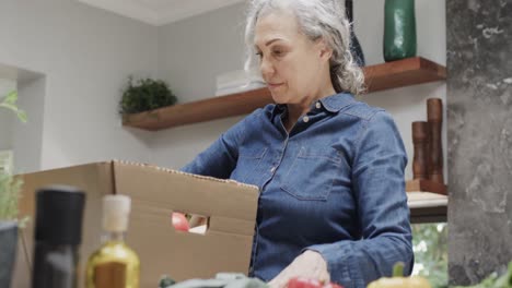 Feliz-Mujer-Caucásica-Mayor-Desempacando-Caja-De-Verduras-Y-Frutas-En-La-Cocina,-Cámara-Lenta