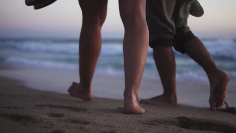 slow motion low angle handheld shot of a couple in love walking at dusk on a beautiful sandy beach in front of the sea with calm waves