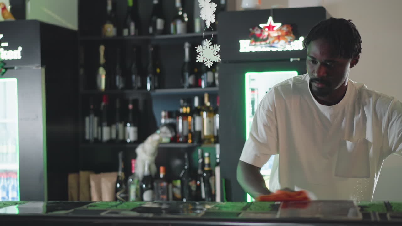 Bartender arranging drinks and glasses behind bar counter in trendy pub. Shelves stocked with bottles and festive decorations, including snowflakes. A rabbit is placed on the counter near bartender