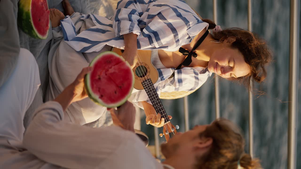 Vertical video of a happy couple on a date. On the pier near the sea, the guy cuts a watermelon, the girl plays the ukulele. Guy and girl on a picnic on the pier near the sea